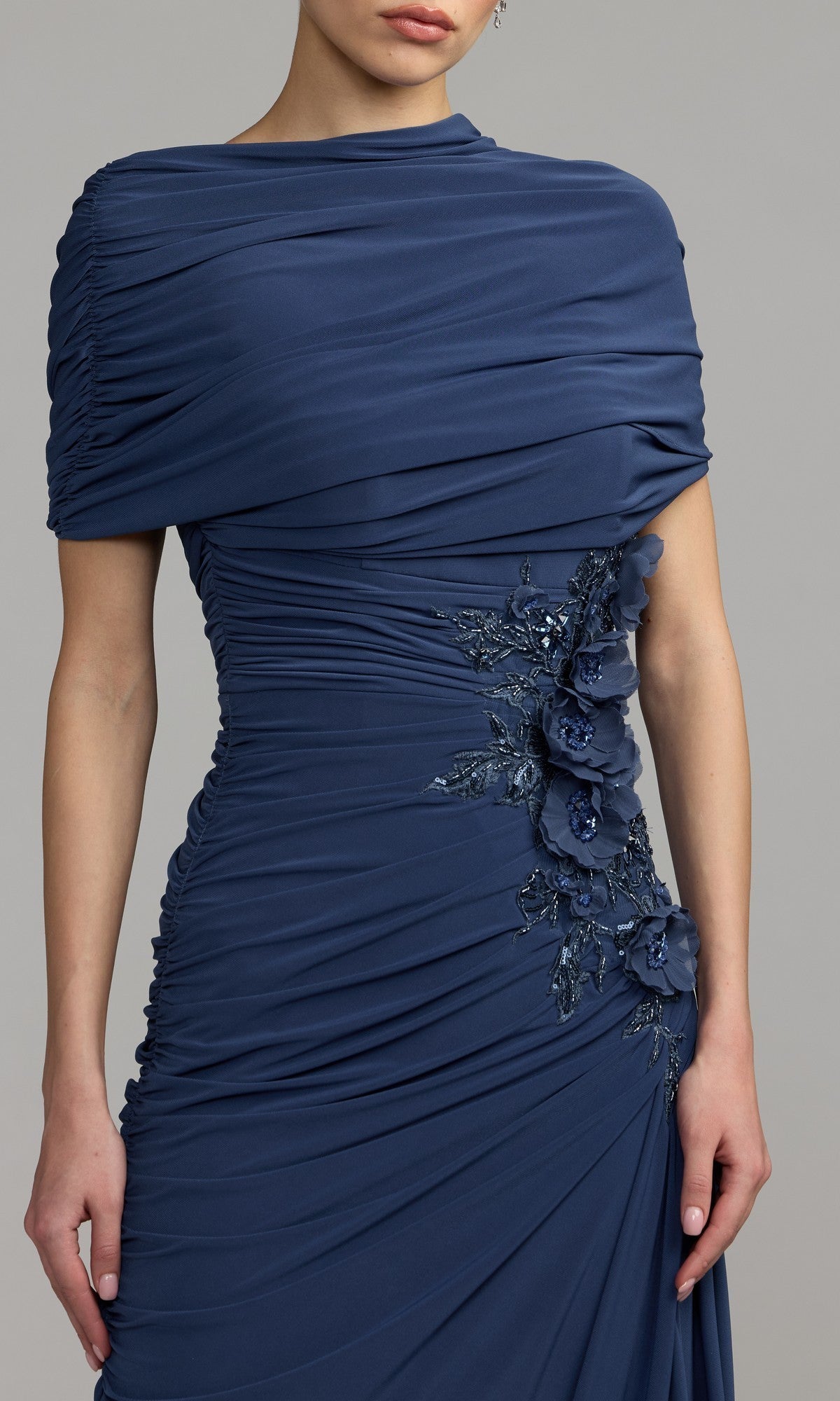 A woman is photographed from the shoulders to mid-thigh, modeling the Mac Duggal 13244 Long Formal Dress—a dark blue ruched gown featuring floral embellishments on the side—against a plain backdrop.